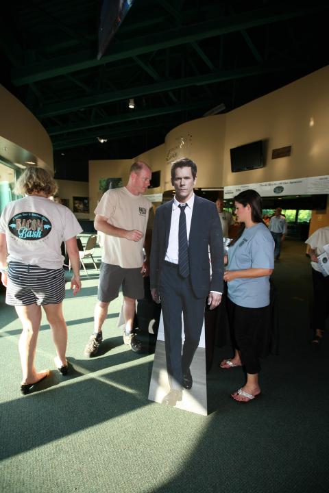 People gather around a life-sized cardboard cutout of a man in a suit inside a well-lit indoor space.