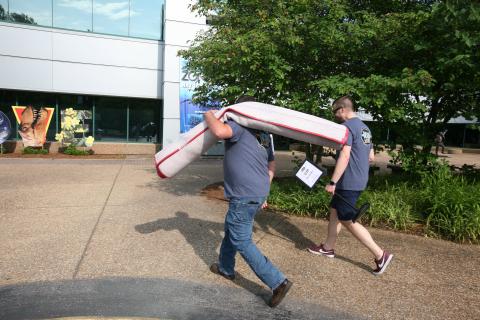 Two men carry a rolled-up rug across a paved area near a modern building with trees in the background