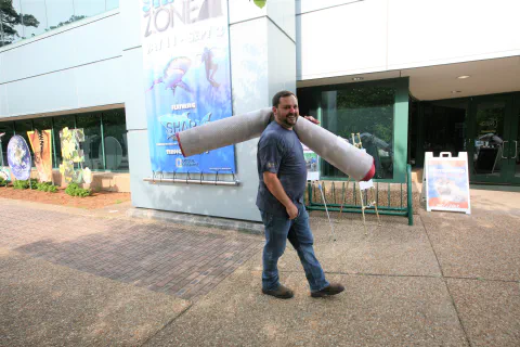 Man carrying large cylindrical object across paved area in front of modern building with colorful signage.