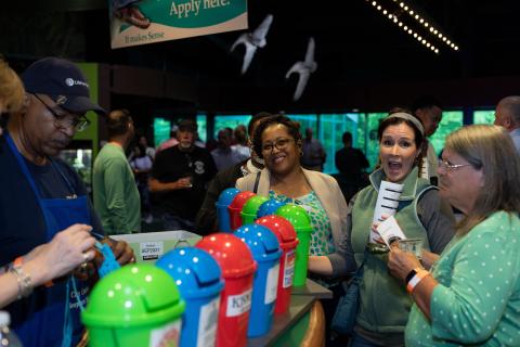 Group of people interacting with colorful recycling bins at an event with birds flying overhead.