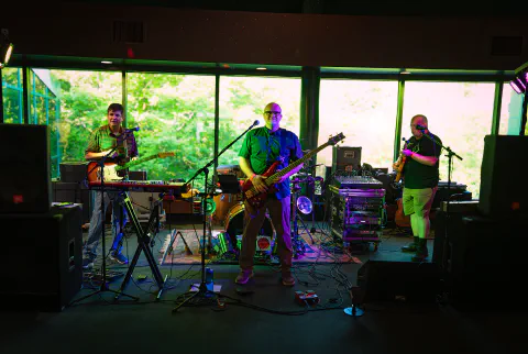 Four musicians perform on stage with guitars, keyboards, and drums, bathed in colorful stage lights against a green backdrop.